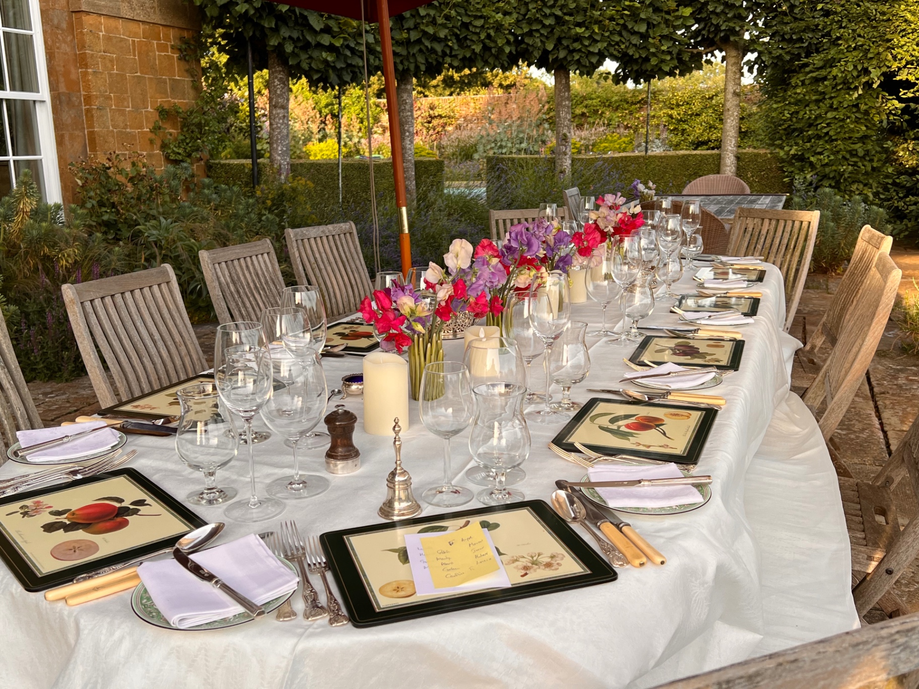 Long linen-dressed dining table set under a walled garden loggia with sweet peas, hurricane candles and botanical placemats — Lovey Chef private dining tablescape in the South West